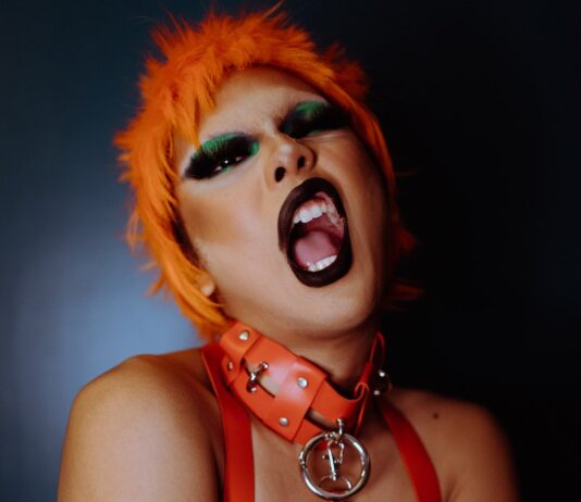 AFTER HOURS POETRY Naughty young ethnic androgynous guy with bright makeup in BDSM collar and wig embracing self and looking at camera with opened mouth